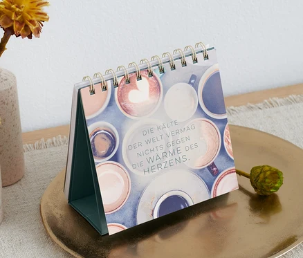 Un livre-présentoir de table « Mach jeden Tag magisch » (en allemand) est posé sur une assiette dorée à côté de vases avec des fleurs.