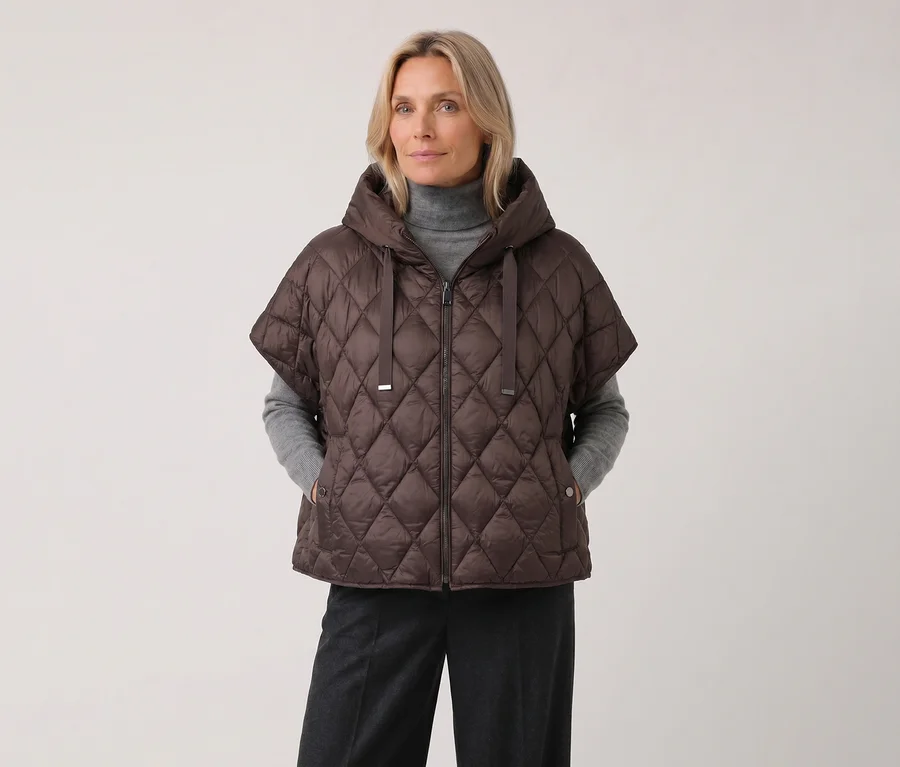 Une femme blonde porte un gilet matelassé Barbara Lebek marron à capuche et manches courtes, un pull à col roulé gris et un pantalon gris foncé.