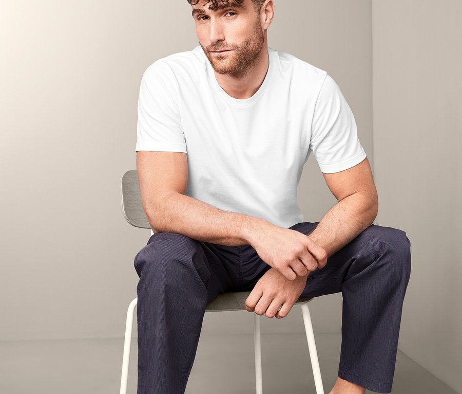 Un homme est assis sur une chaise et porte un t-shirt blanc et un pantalon bleu. Il porte l'un des 2 t-shirts à encolure ronde.