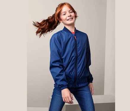 Une jeune fille avec une queue de cheval rousse pose dans une veste style bomber recyclée bleue et un jean.