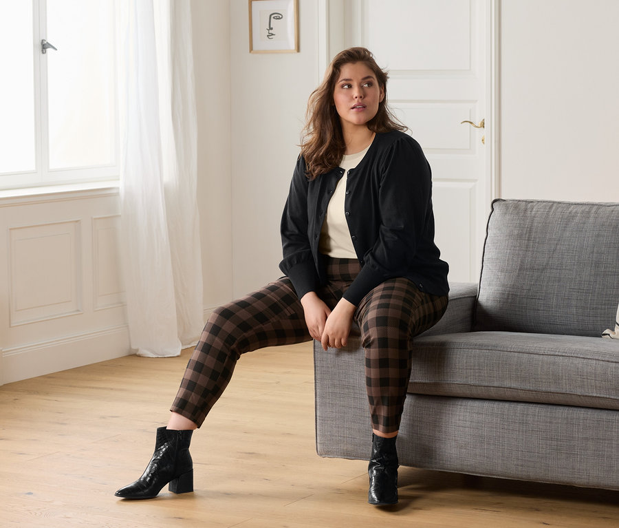 Une femme est assise sur un canapé gris et porte un t-shirt crème, un pantalon stretch à carreaux et une jaquette en maille fine avec des manches bouffantes.