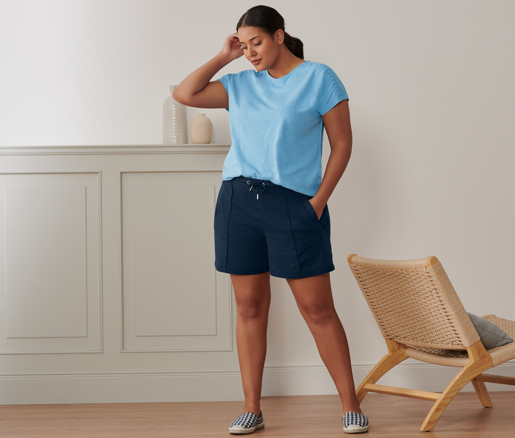 Une femme se tient dans une pièce et porte un t-shirt en lin mélangé bleu clair, un short molletonné et des espadrilles.