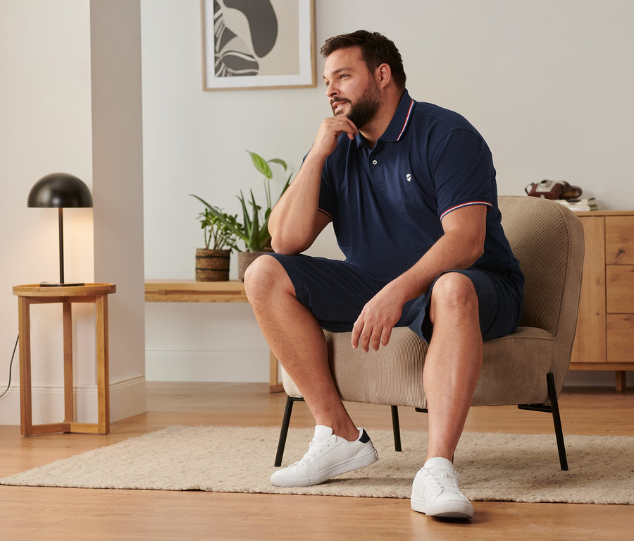 Un homme est assis sur une chaise, portant un short chino bleu marine, des tennis et un polo en piqué bleu marine.