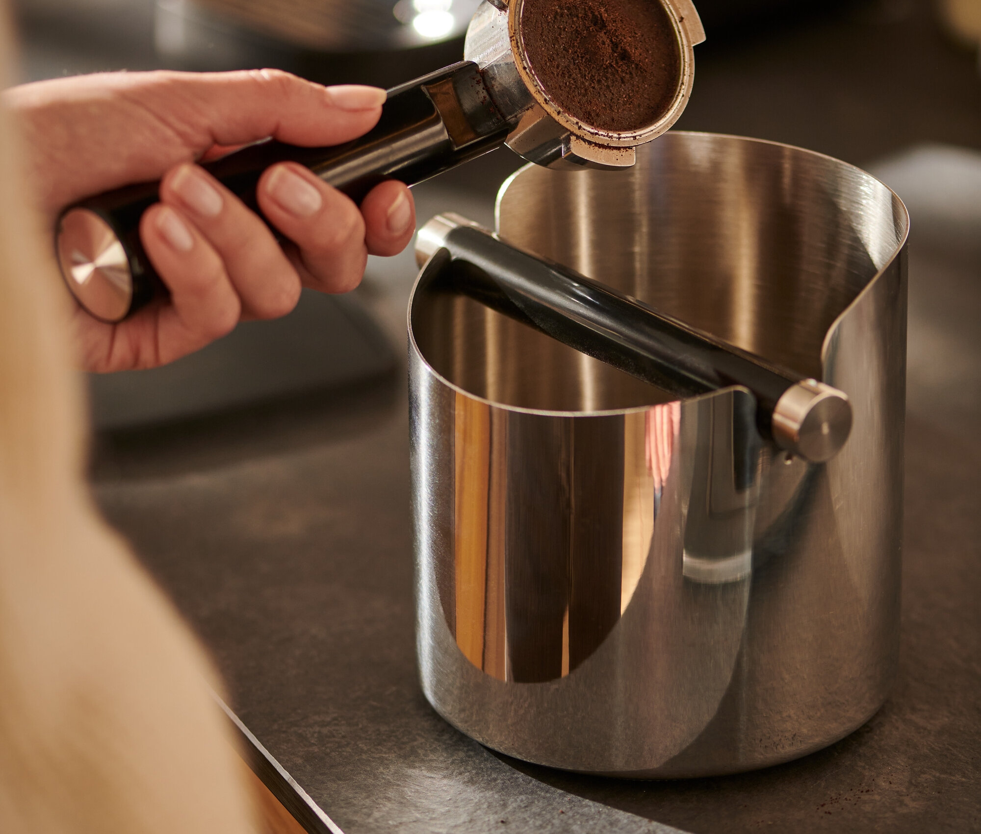 Une main tient un porte-filtre à café au-dessus d'une boîte à marc de café brillante.