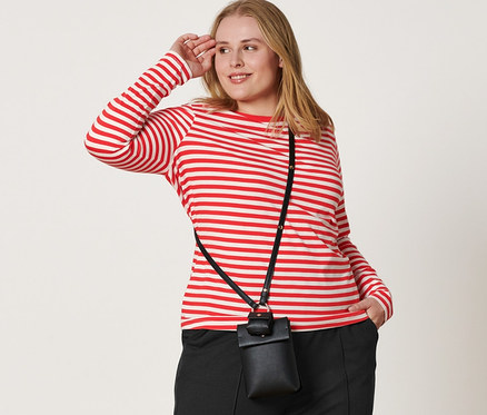 Une femme pose, portant un T-shirt à manches longues rayé rouge et blanc, un pantalon de jogging noir avec une nervure, des baskets en cuir et un mini sac en bandoulière.