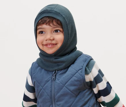 Un garçon souriant porte une cagoule pour tout-petits, un gilet matelassé et un pull rayé.