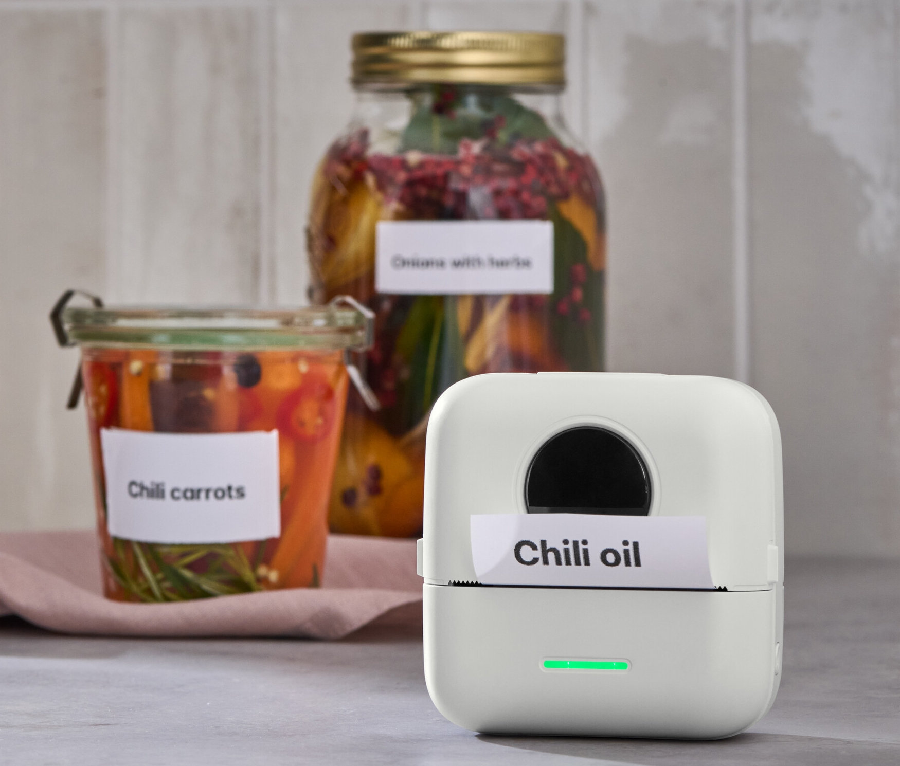 Une mini-imprimante thermique portative imprime une étiquette avec l'inscription Huile de chili. En arrière-plan, bocaux de carottes au chili.