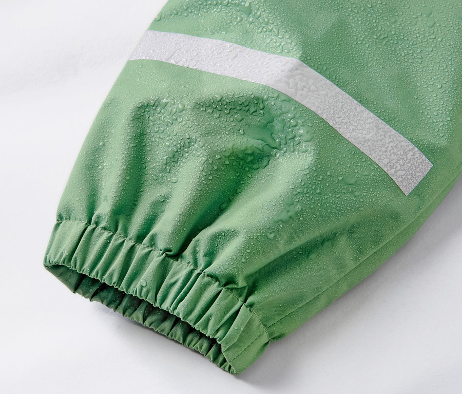 Détail d'un pantalon de pluie pour enfant, vert gazon, avec une bande réfléchissante et des gouttes d'eau.