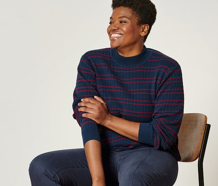 Une femme assise sur une chaise porte un pull en maille fine bleu rouge, un pantalon twill bleu foncé et des loafers en cuir.