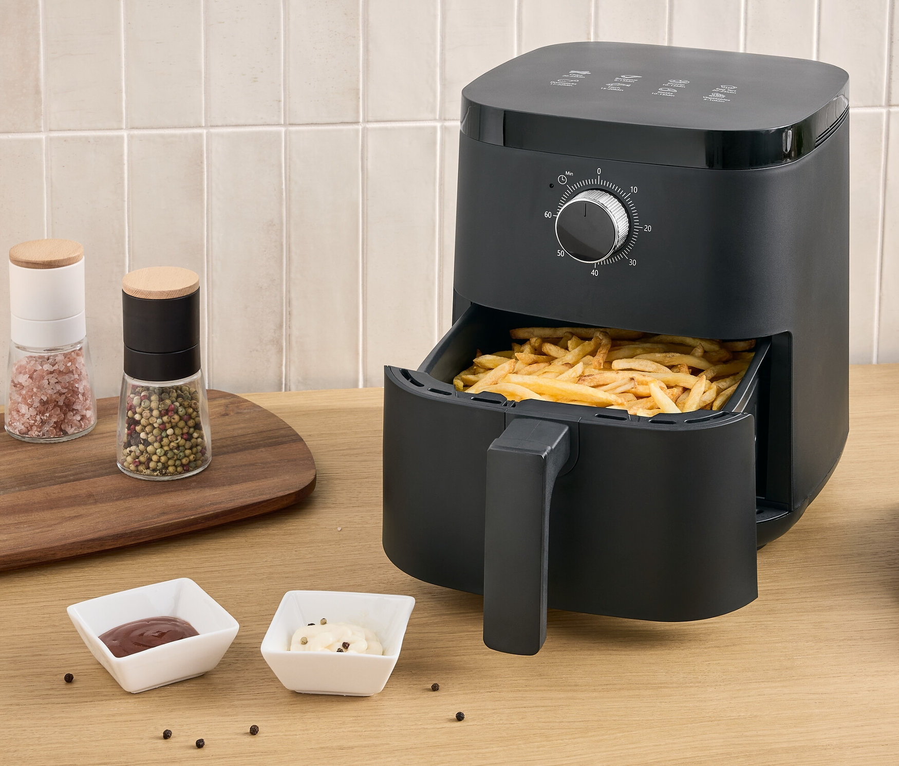 Une friteuse à air chaud noire avec des frites dans le tiroir se trouve sur une table à côté d'une assiette de frites et de deux sauces.