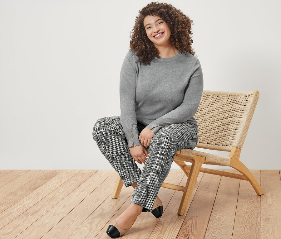 Une femme souriante est assise sur une chaise et porte un pull en maille fine gris chiné et un pantalon stretch imprimé bleu marine.
