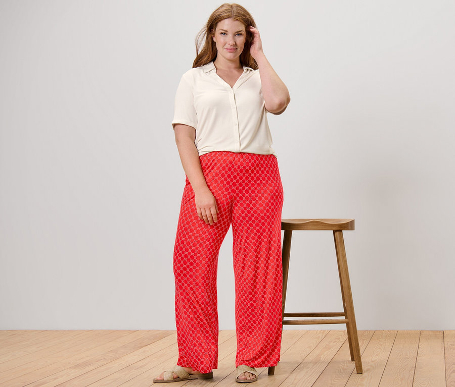 Une femme pose en pantalon palazzo imprimé rouge et blouse blanche avec patte de boutonnage.