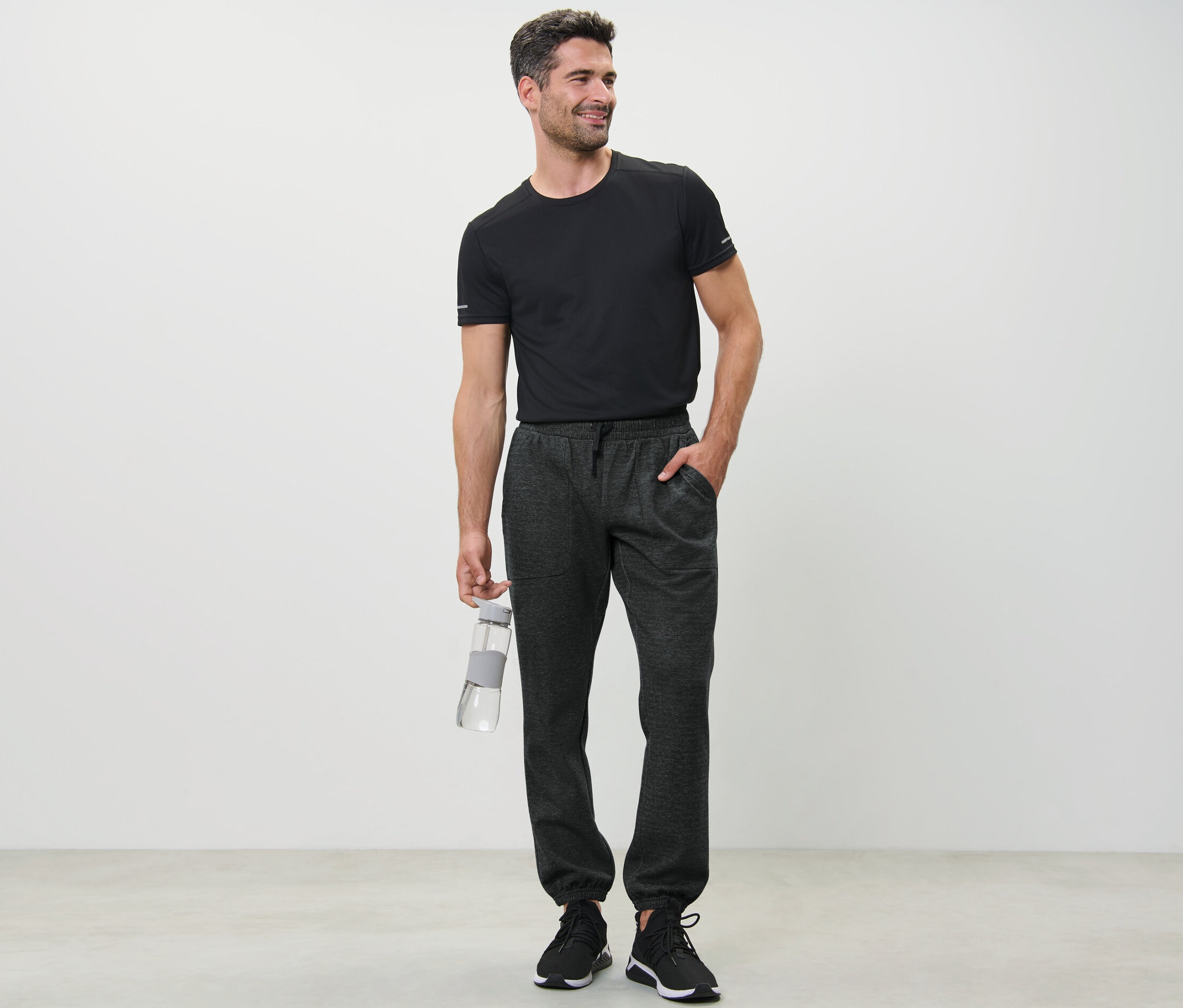 Un homme en t-shirt noir tenant une bouteille d'eau et portant un pantalon de survêtement.