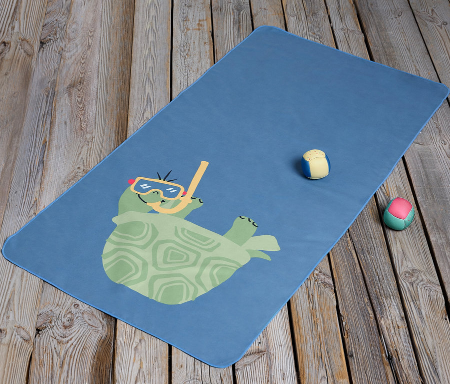 Une serviette microfibre bleue pour enfants avec une tortue de dessin animé portant des lunettes et un tuba est posée sur un plancher en bois. Deux petites balles sont à côté de la serviette.