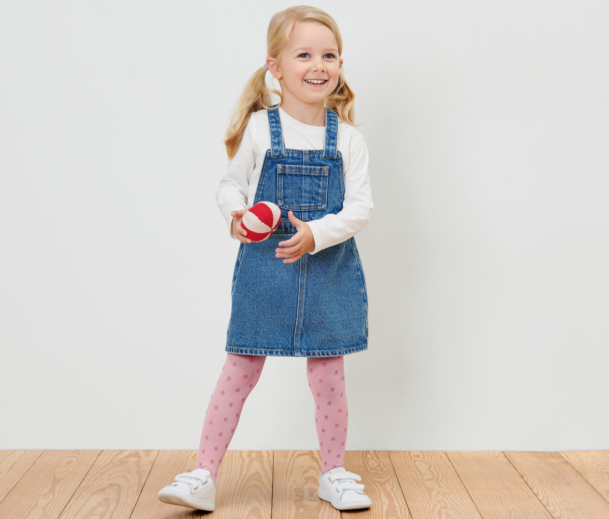 Une fillette blonde souriante en robe en jean tient une balle rouge et blanche et porte 2 collants fins pour enfant.