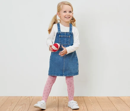 Une fillette blonde souriante en robe en jean tient une balle rouge et blanche et porte 2 collants fins pour enfant.