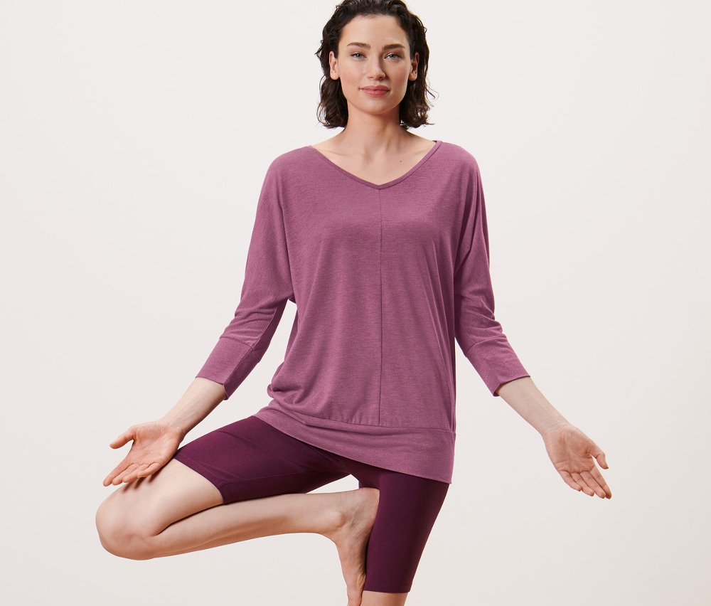 Femme faisant la posture de l'arbre sur un tapis de yoga. Elle porte un t-shirt de yoga mauve.