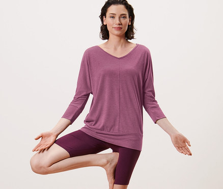 Femme faisant la posture de l'arbre sur un tapis de yoga. Elle porte un t-shirt de yoga mauve.