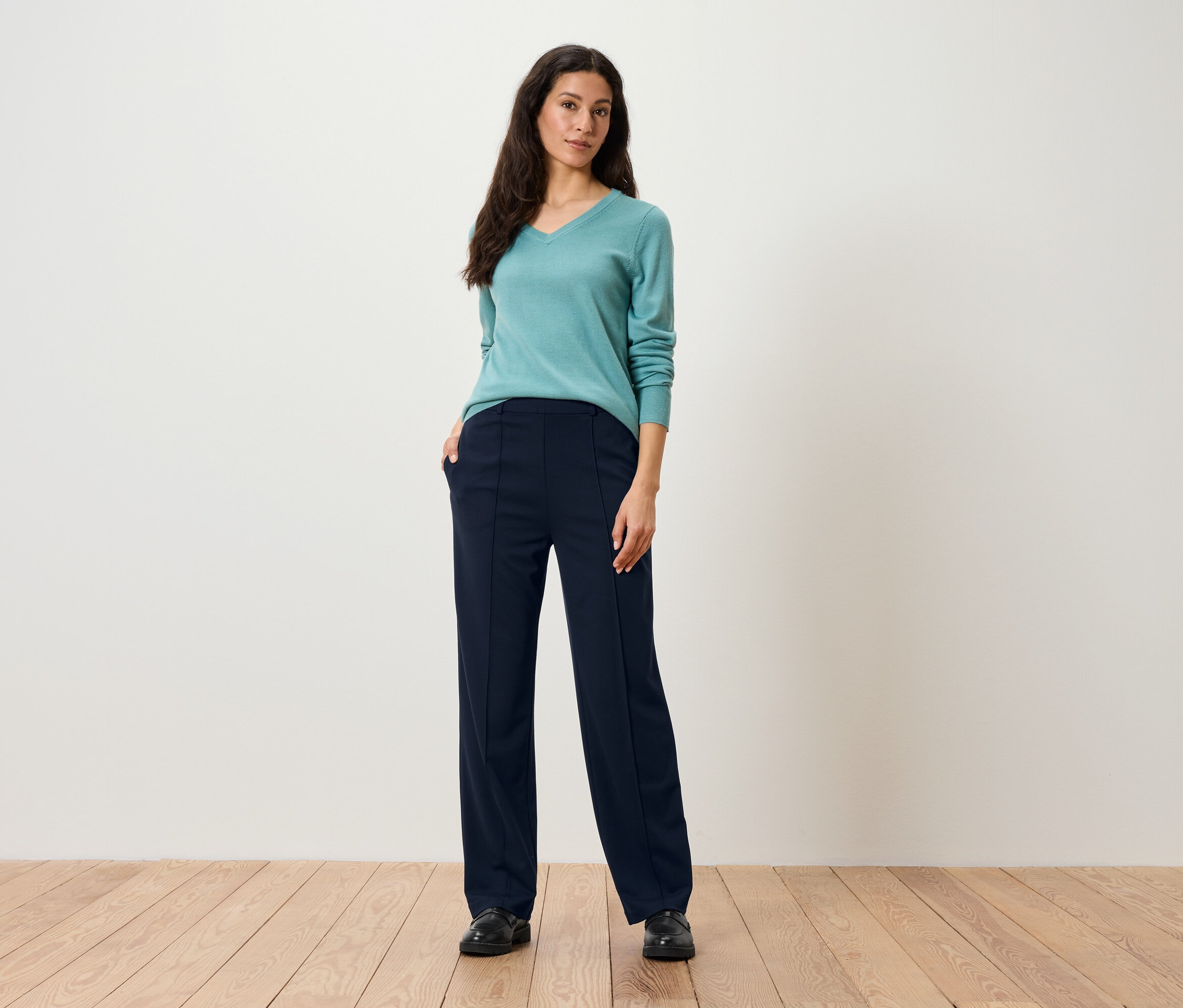 Une femme se tient debout et pose en pantalon bleu et pull turquoise.