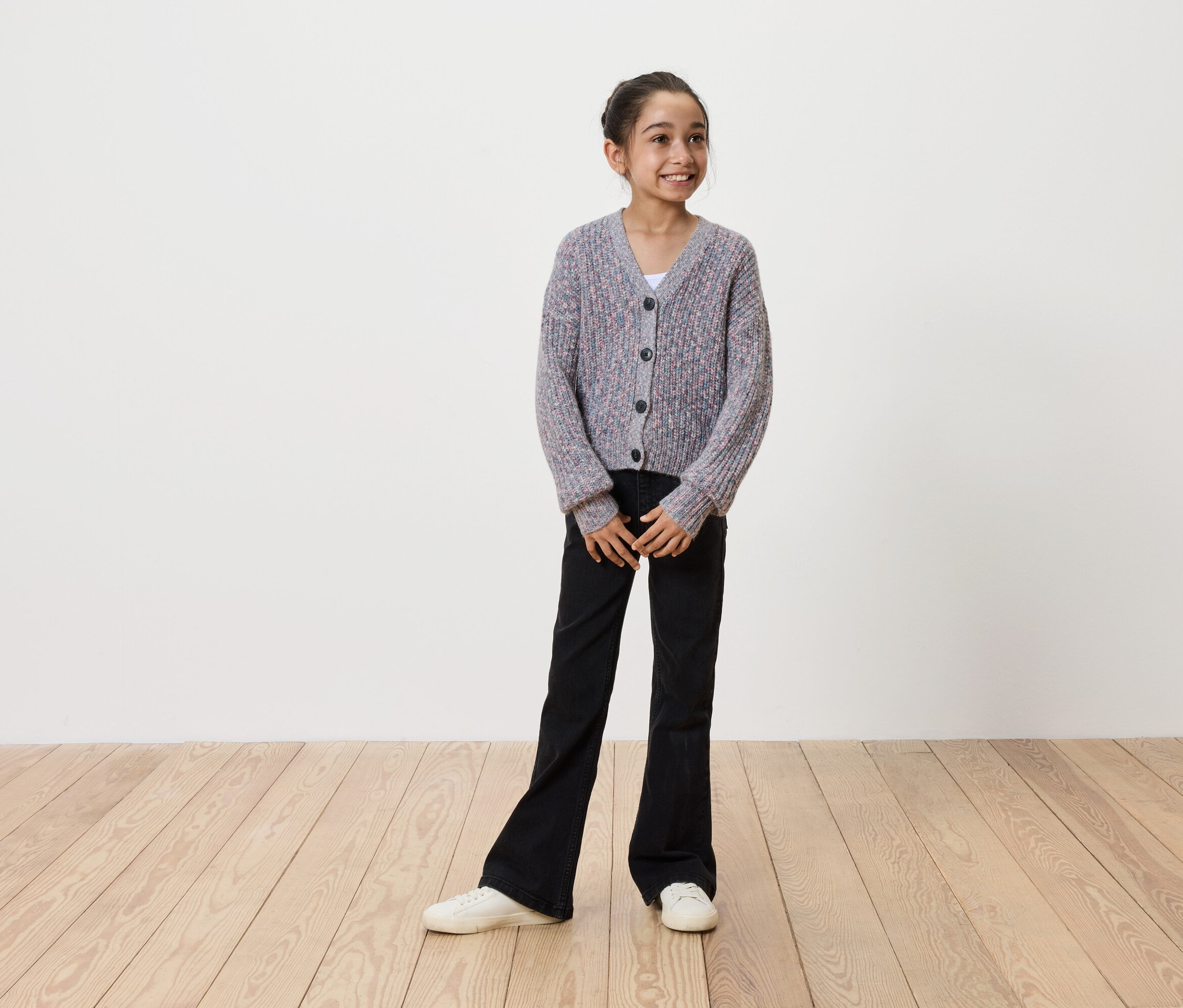 Une fillette souriante portant un cardigan en tricot gris boutonné, un pantalon noir évasé et des baskets blanches, se tient sur un plancher en bois devant un mur blanc.