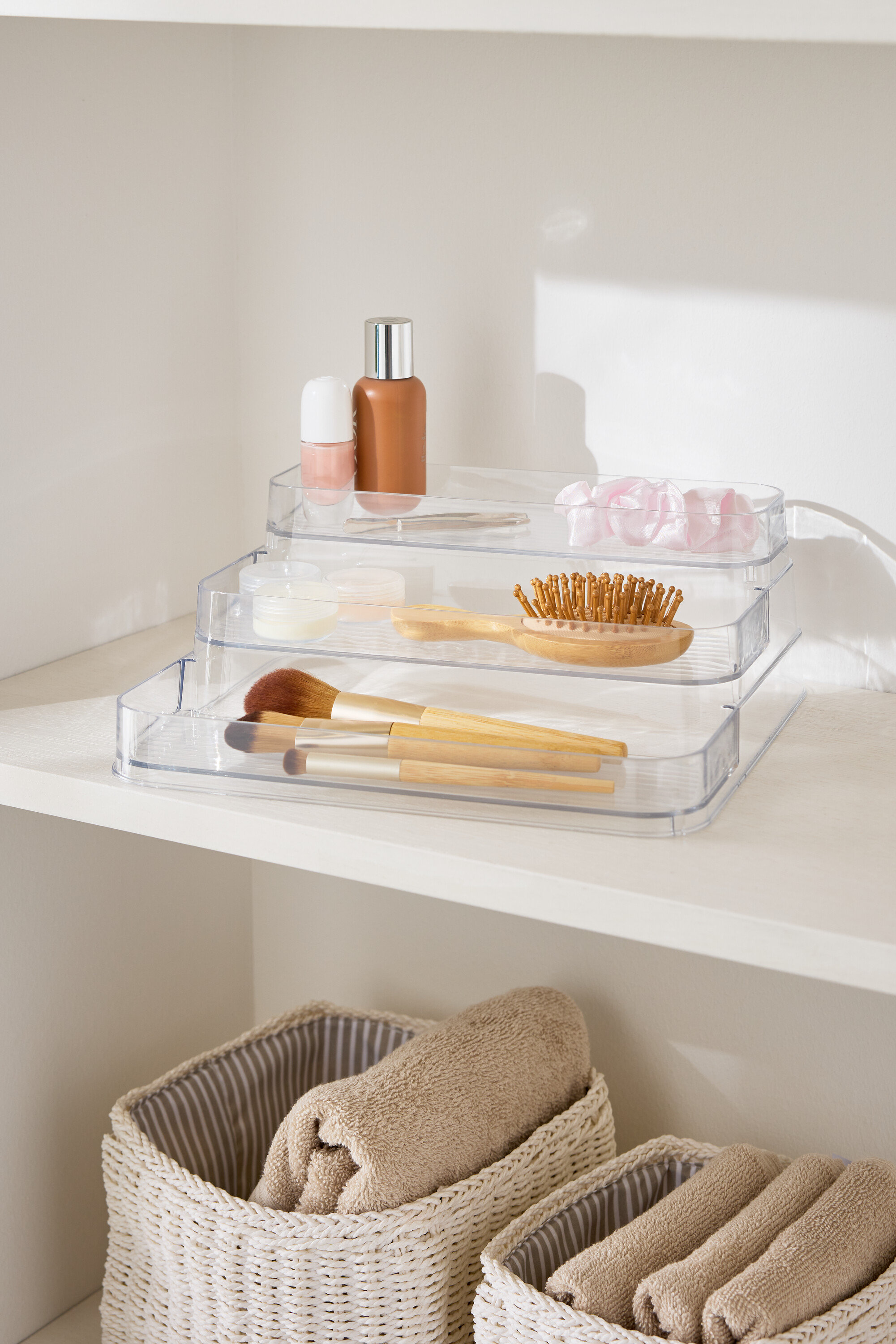 Organisateur de maquillage transparent à trois niveaux sur une étagère blanche à côté de paniers avec des serviettes.