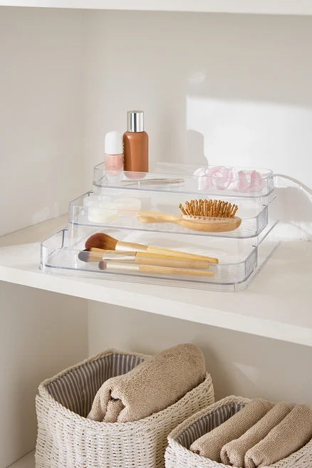 Organisateur de maquillage transparent à trois niveaux sur une étagère blanche à côté de paniers avec des serviettes.