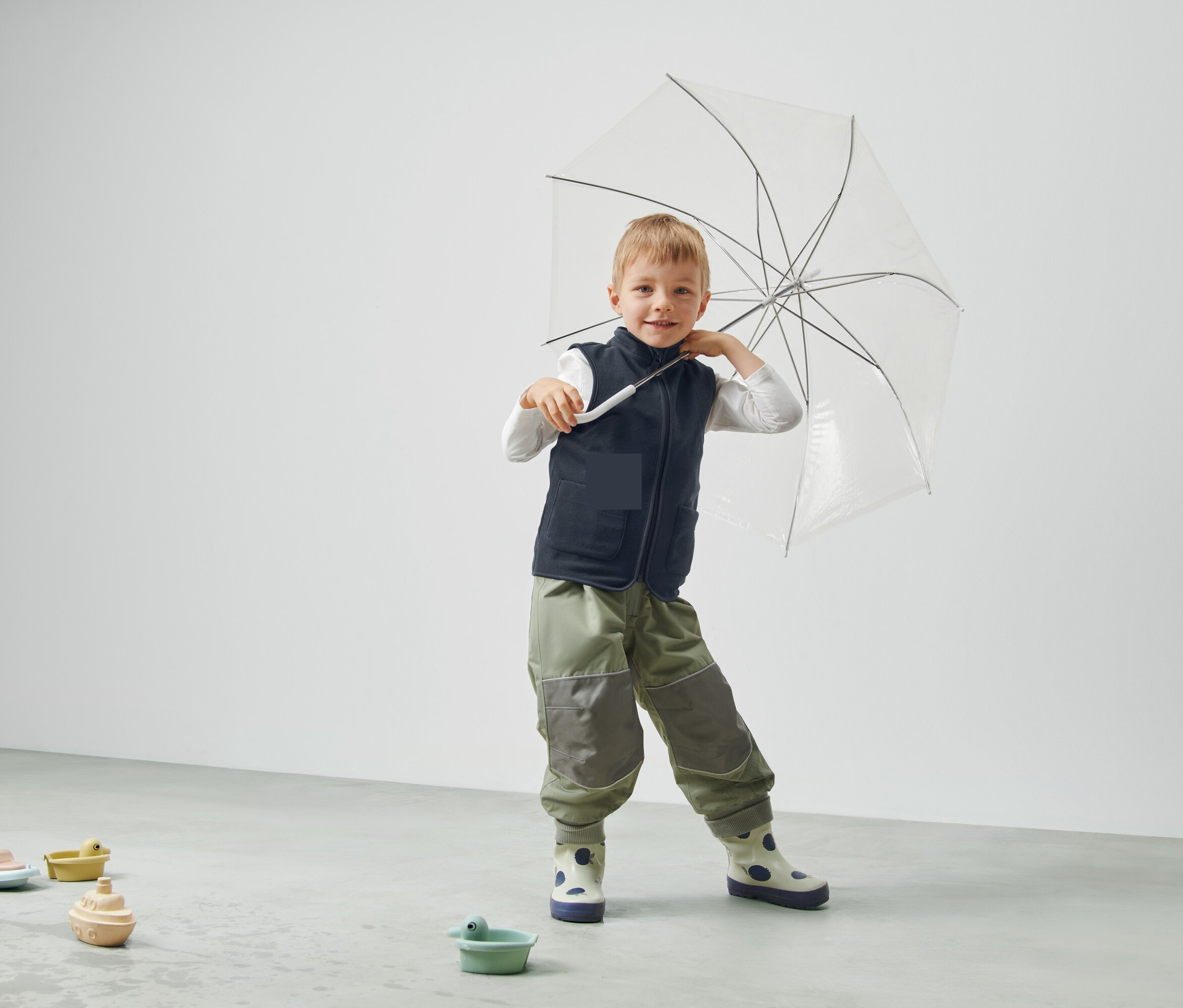 Un garçon tient un parapluie transparent, porte un gilet et un pantalon et se tient dans une pièce.
