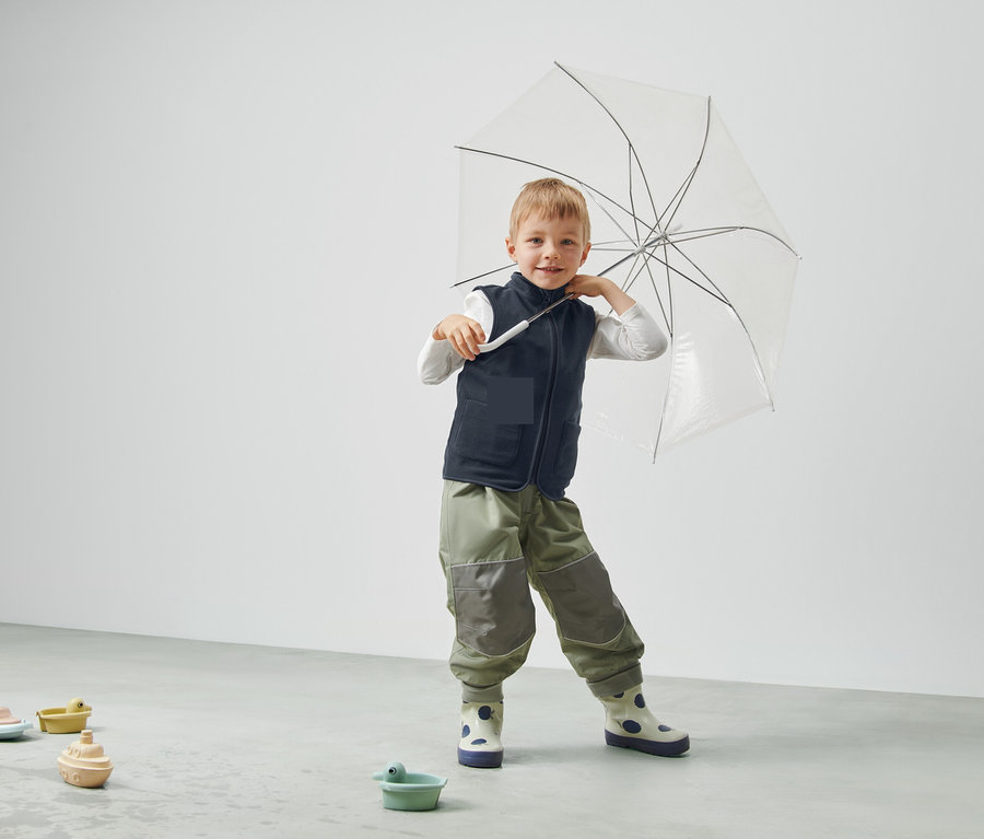 Un garçon tient un parapluie transparent, porte un gilet et un pantalon et se tient dans une pièce.