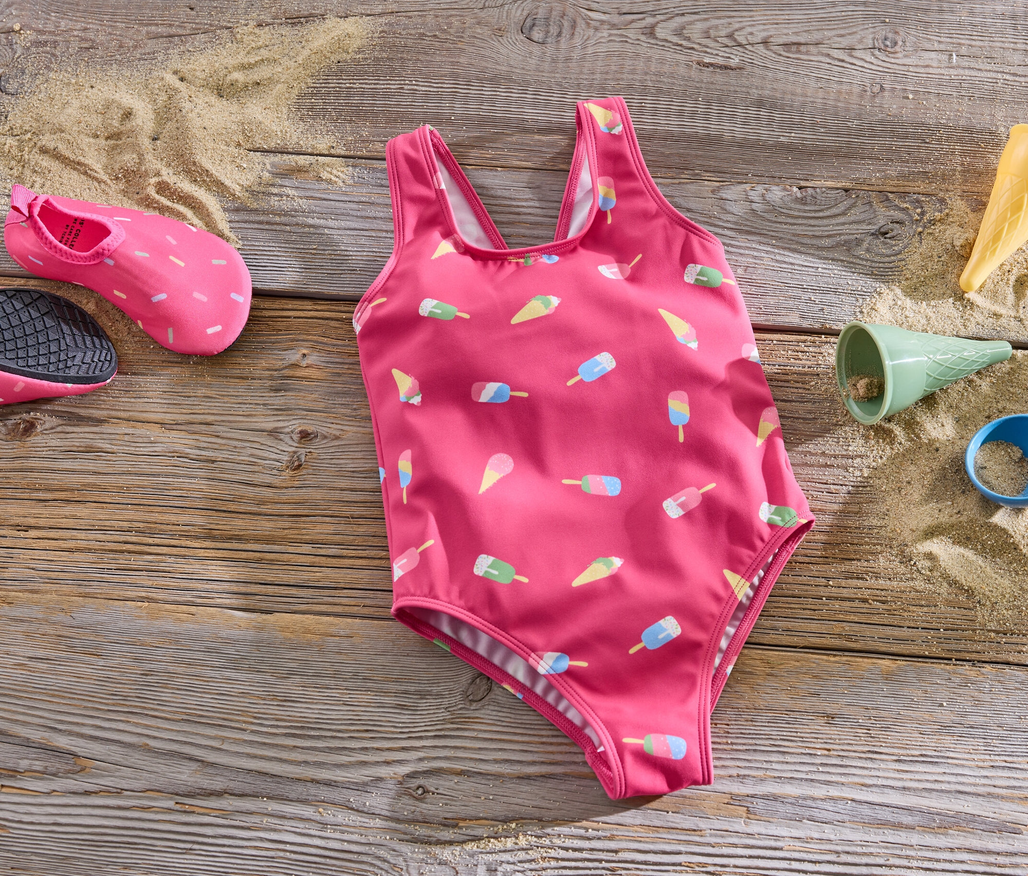 Un maillot de bain pour enfant rose avec des motifs de glace posé sur une surface en bois à côté de chaussures d'eau et de jouets de plage.
