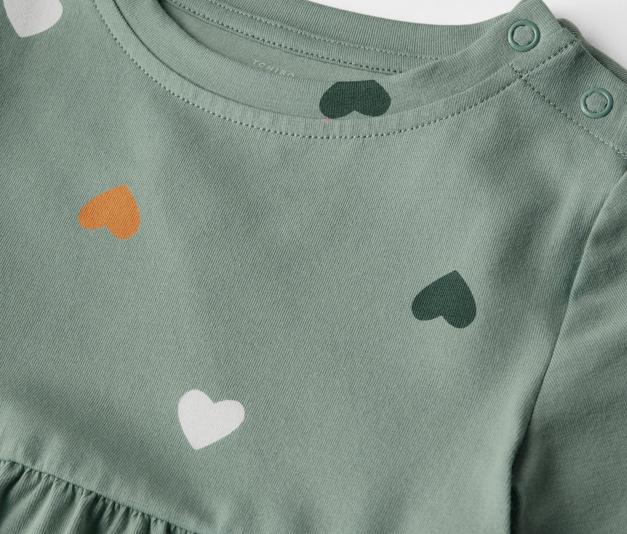 Gros plan d'une robe en jersey vert clair pour enfants avec un imprimé de cœurs et des boutons sur l'épaule.