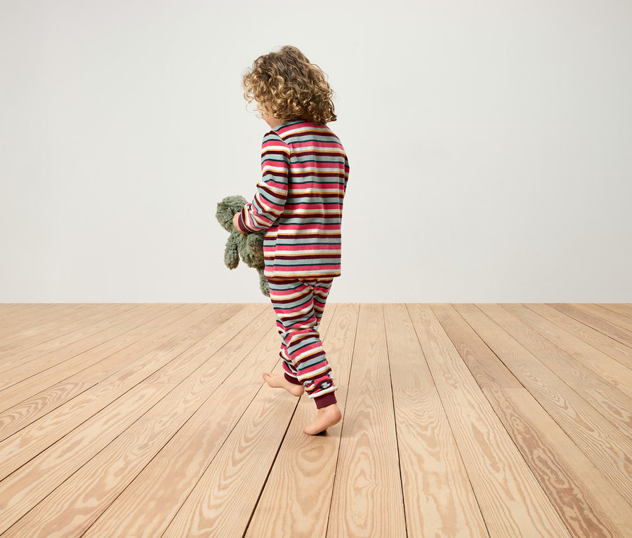 Un enfant aux cheveux bouclés marche pieds nus sur un plancher en bois en pyjama rayé et tient un ours en peluche.