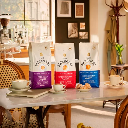 Trois paquets de café Molinari sont posés sur une table à côté de tasses et d'assiettes de pâtisseries.