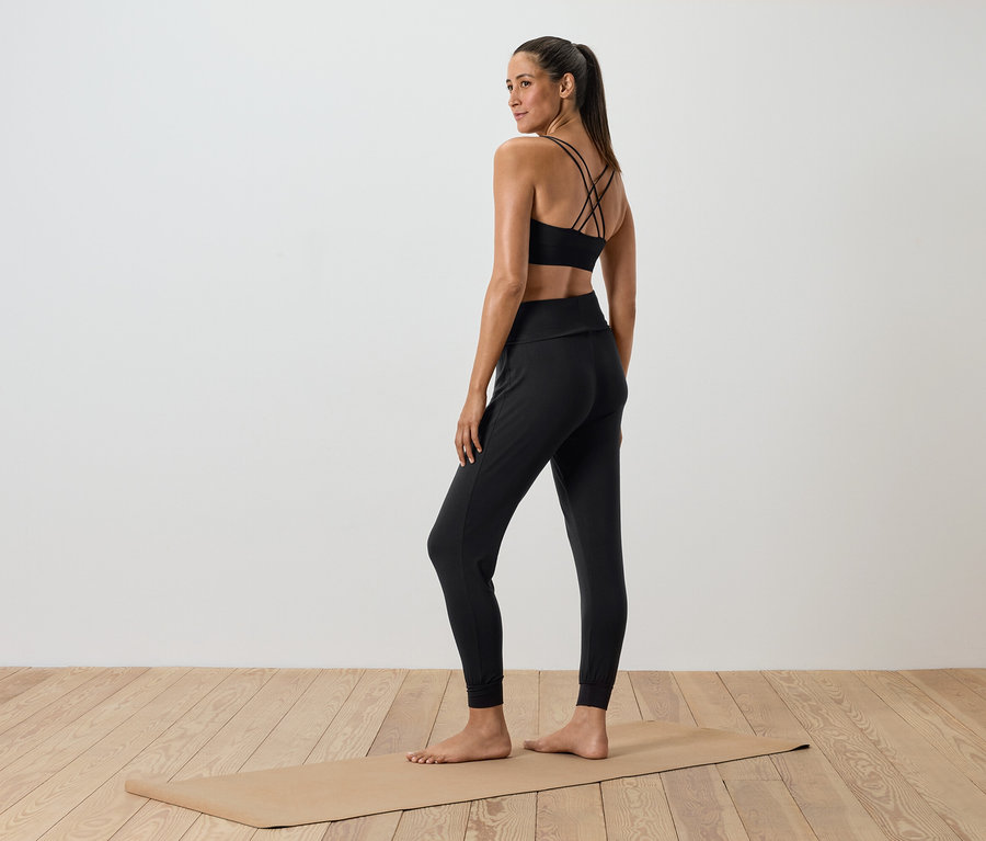 Femme en tenue de sport noire debout sur un tapis de yoga.