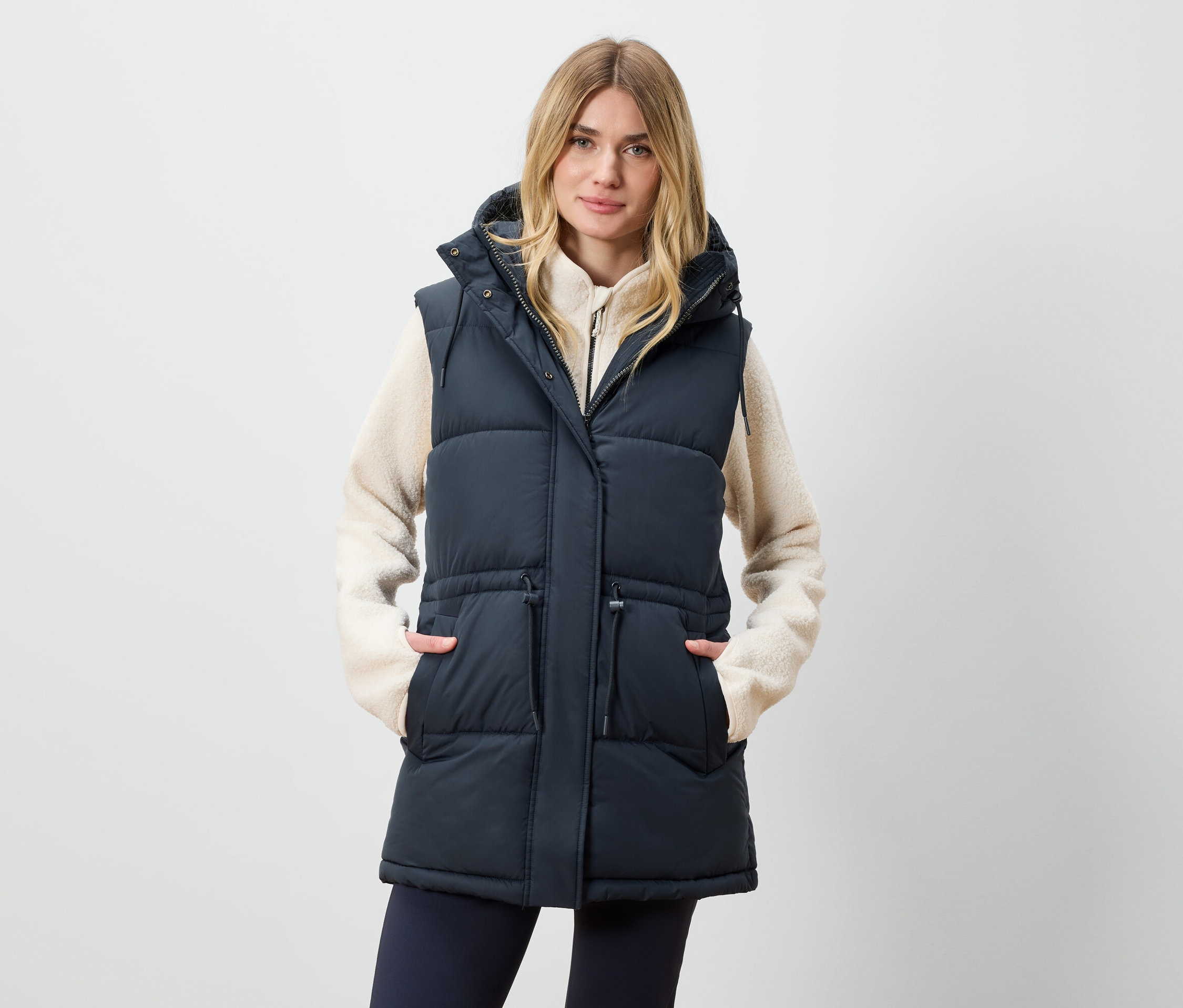 Une femme porte un long gilet matelassé à capuche et a les mains dans les poches.