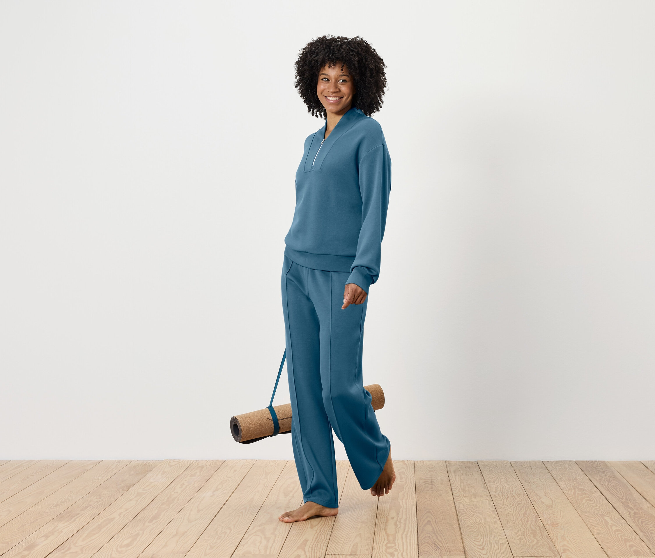 Femme souriante avec une coiffure afro marchant pieds nus et portant un tapis de yoga, vêtue d'un ensemble de survêtement bleu.