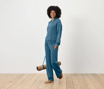 Femme souriante avec une coiffure afro marchant pieds nus et portant un tapis de yoga, vêtue d'un ensemble de survêtement bleu.