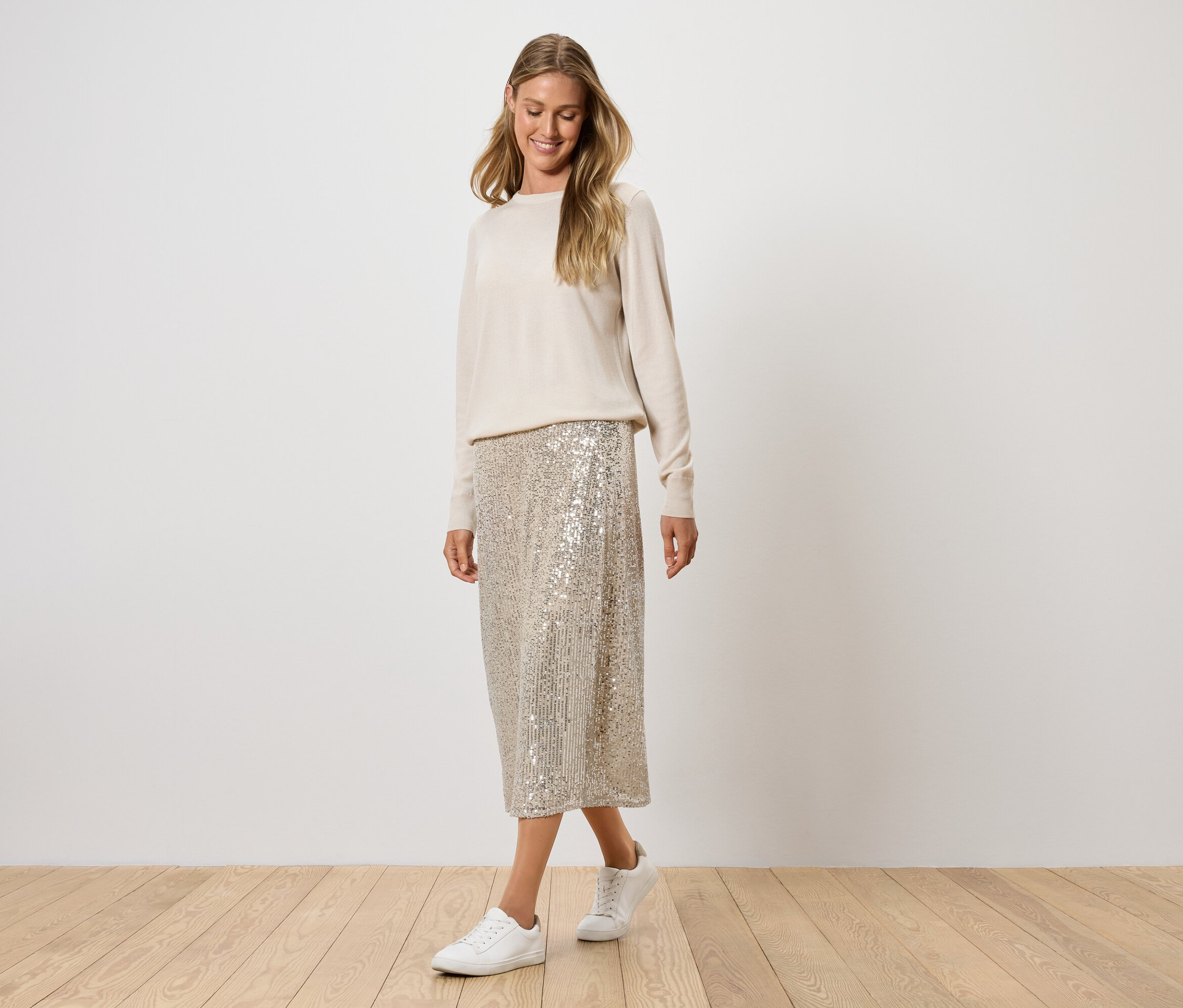 Femme posant en pull crème, jupe à paillettes et baskets blanches.