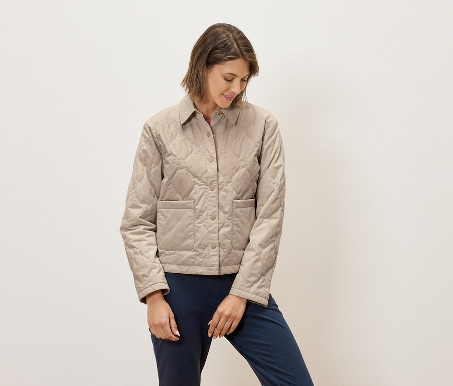 Une femme portant une veste matelassée beige et un pantalon bleu se tient devant un fond blanc.