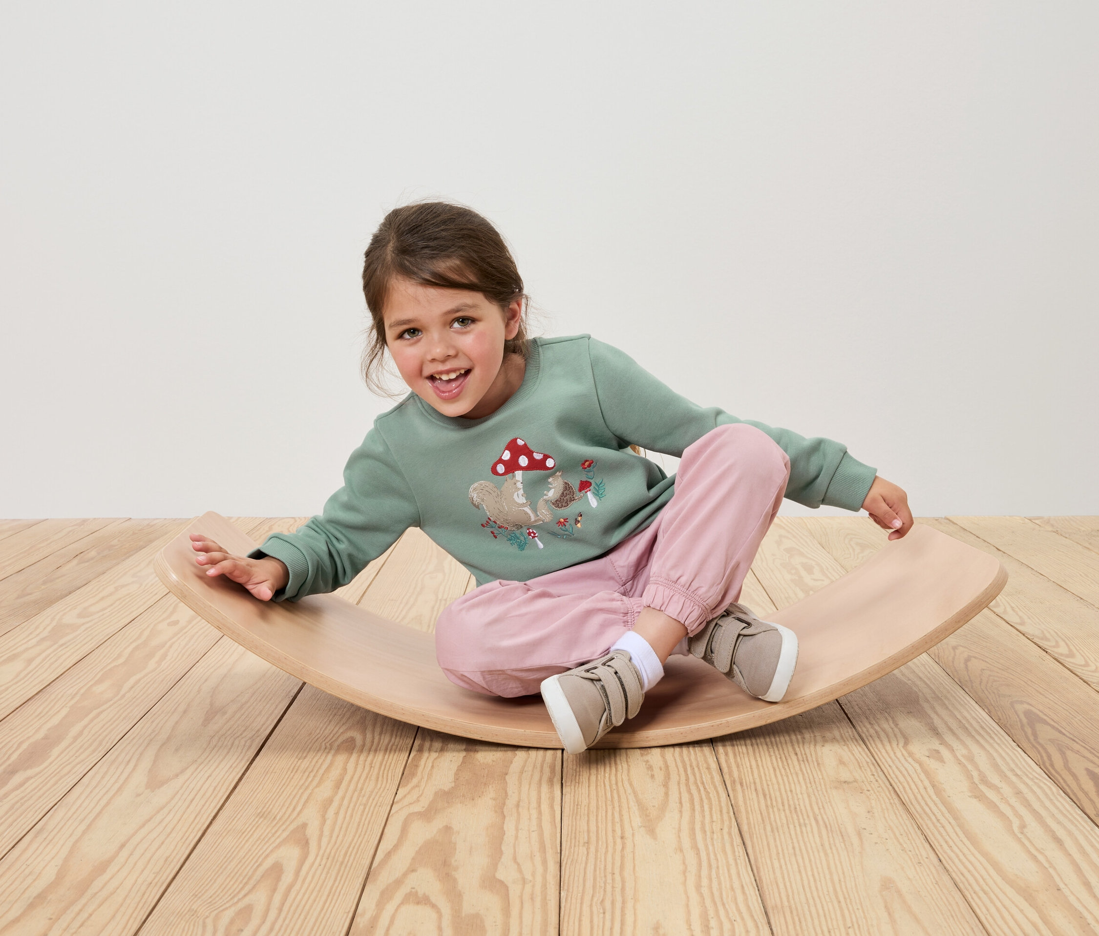 Une fille est assise sur une planche d'équilibre en bois, portant un sweat-shirt vert avec un motif d'écureuil et de champignon, un pantalon rose et des chaussures beiges.