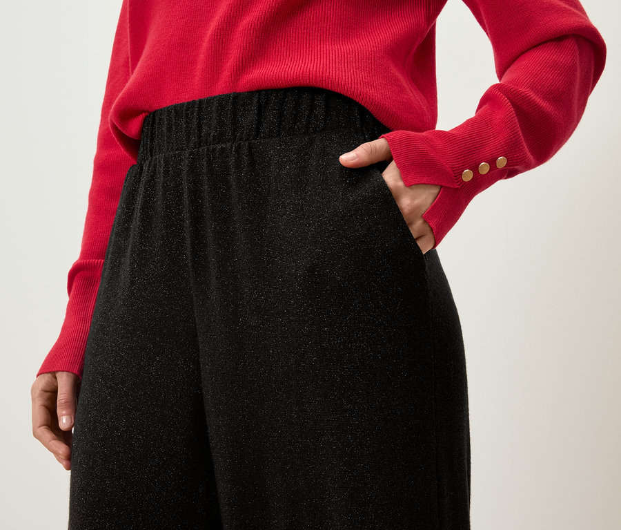 Vue partielle d'une femme portant un pull rouge avec la main dans la poche d'un pantalon noir scintillant.