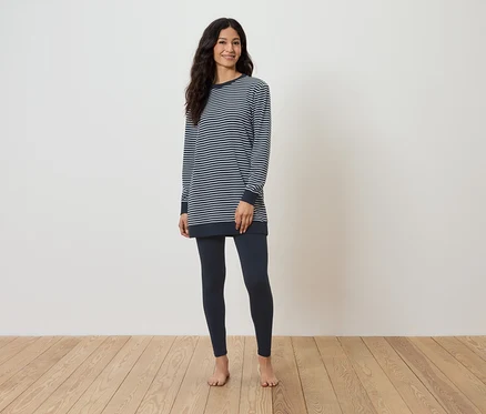 Une femme debout pieds nus sur un plancher en bois, porte un t-shirt rayé à manches longues et un legging bleu foncé.