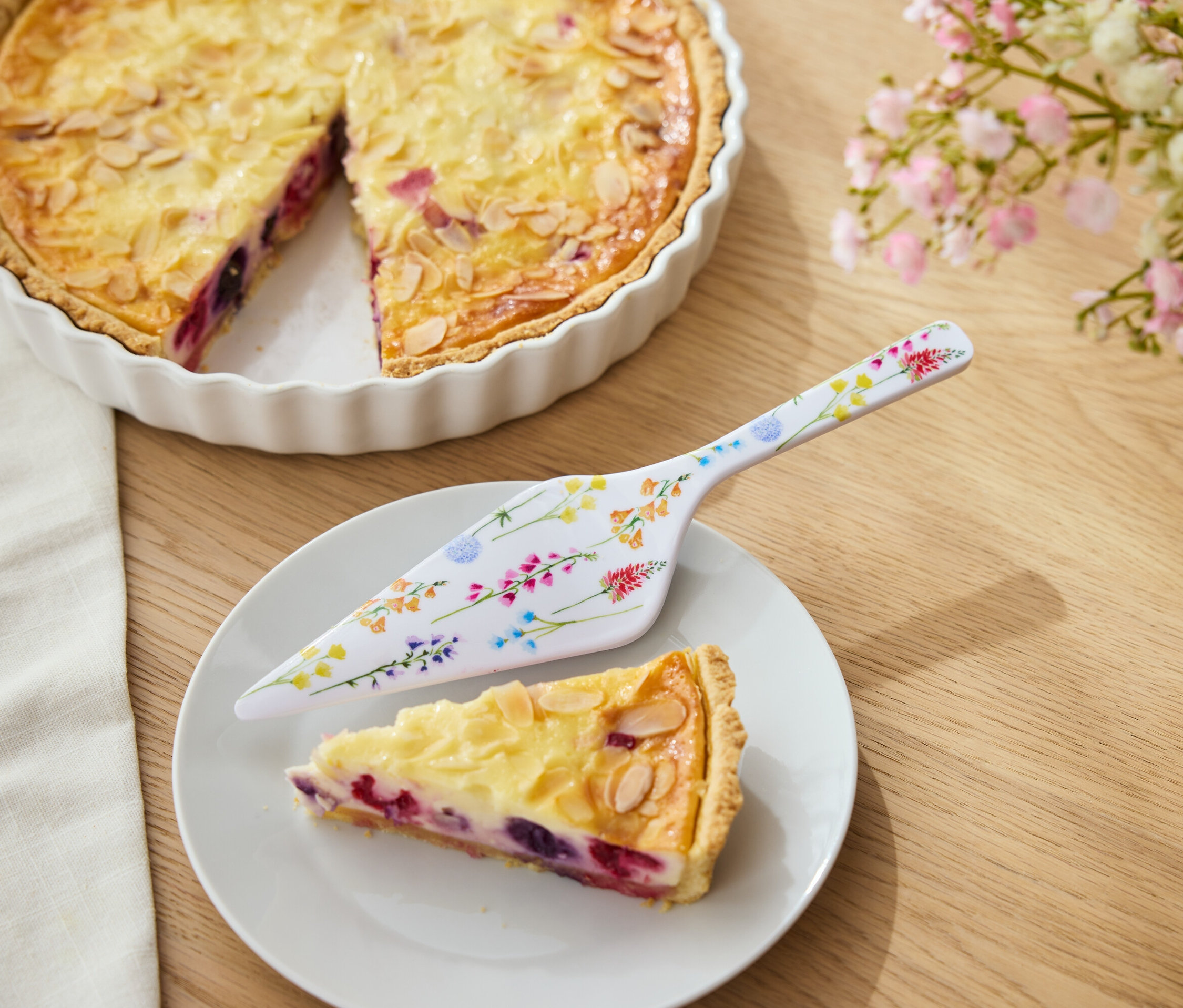 Tarte aux fruits et aux amandes dans un moule blanc, une part de tarte sur une assiette et une pelle à tarte à fleurs.