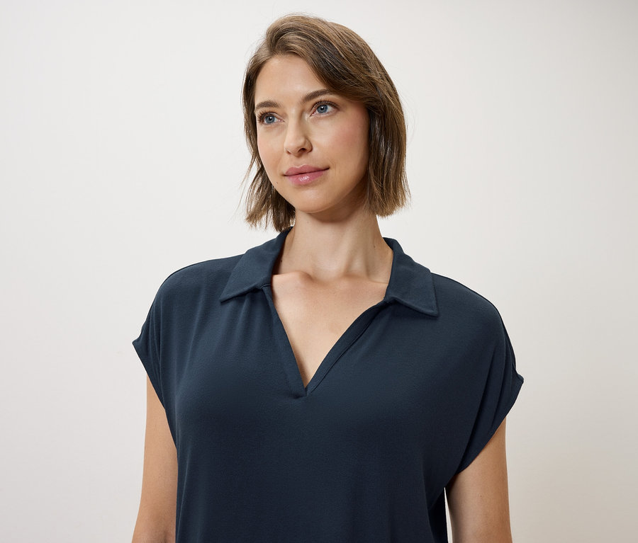 Une femme aux cheveux bruns et aux yeux bleus porte une robe polo bleu foncé.