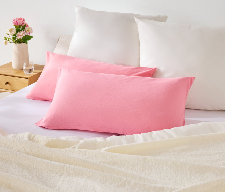 Sur un lit, il y a deux oreillers roses et deux oreillers blancs. Sur la table de chevet, il y a un vase avec des fleurs.