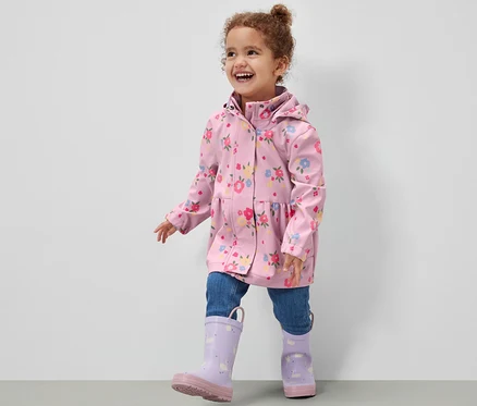 Fillette souriante portant une veste en softshell pour enfant à motif floral, un jean et des bottes de pluie violettes marchant sur un sol gris devant un mur blanc.