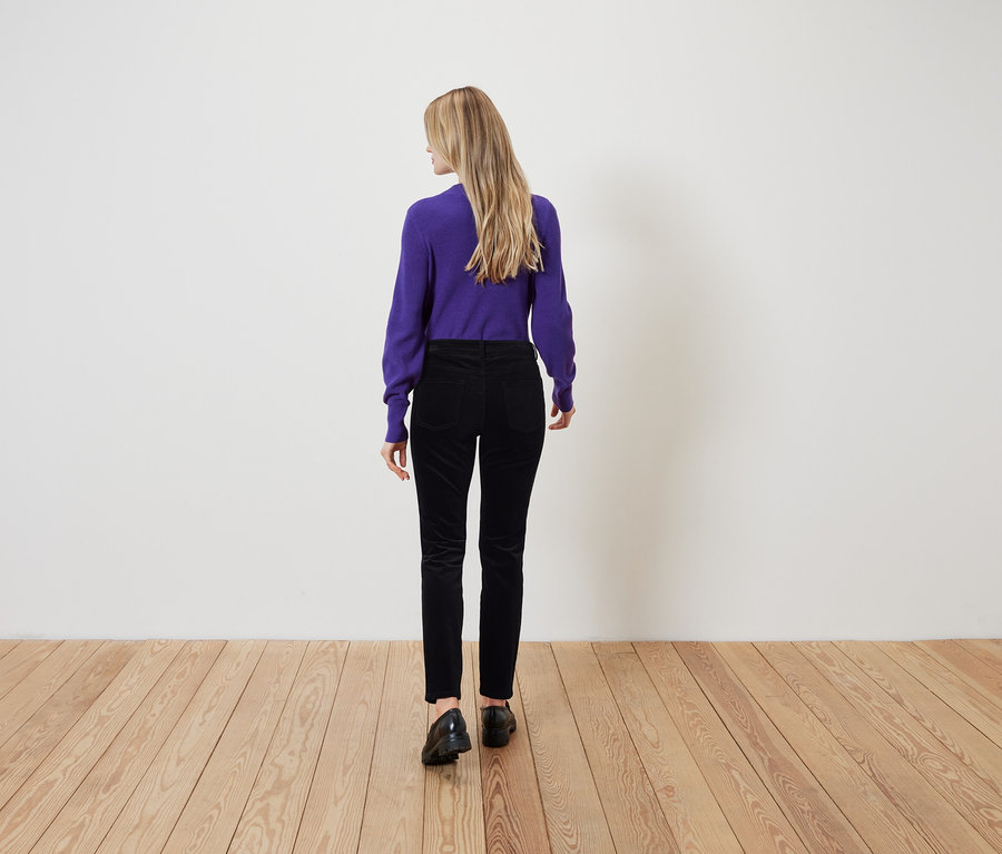 Une femme aux cheveux blonds se tient dos à la caméra portant un pull violet et un pantalon noir.