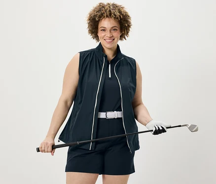 Femme tenant un club de golf portant un top fonctionnel bleu foncé, un short technique en softshell et une veste en softshell.