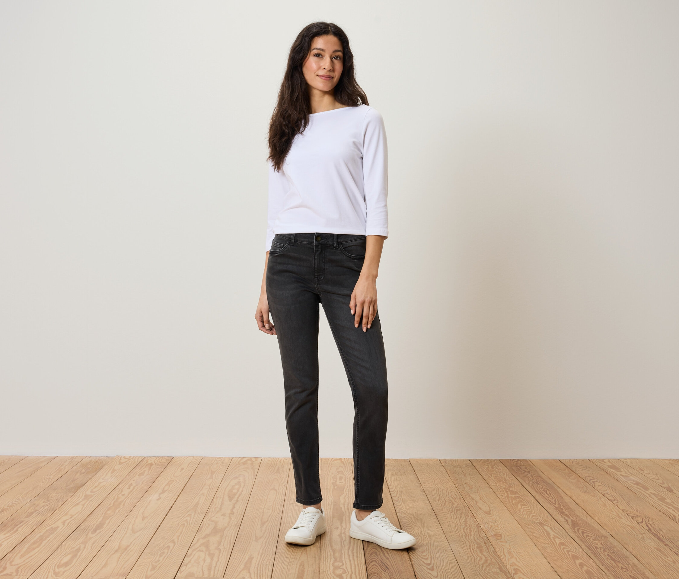 Une femme se tient sur un plancher en bois, portant un t-shirt blanc à manches trois-quarts, un jean gris et des baskets blanches.