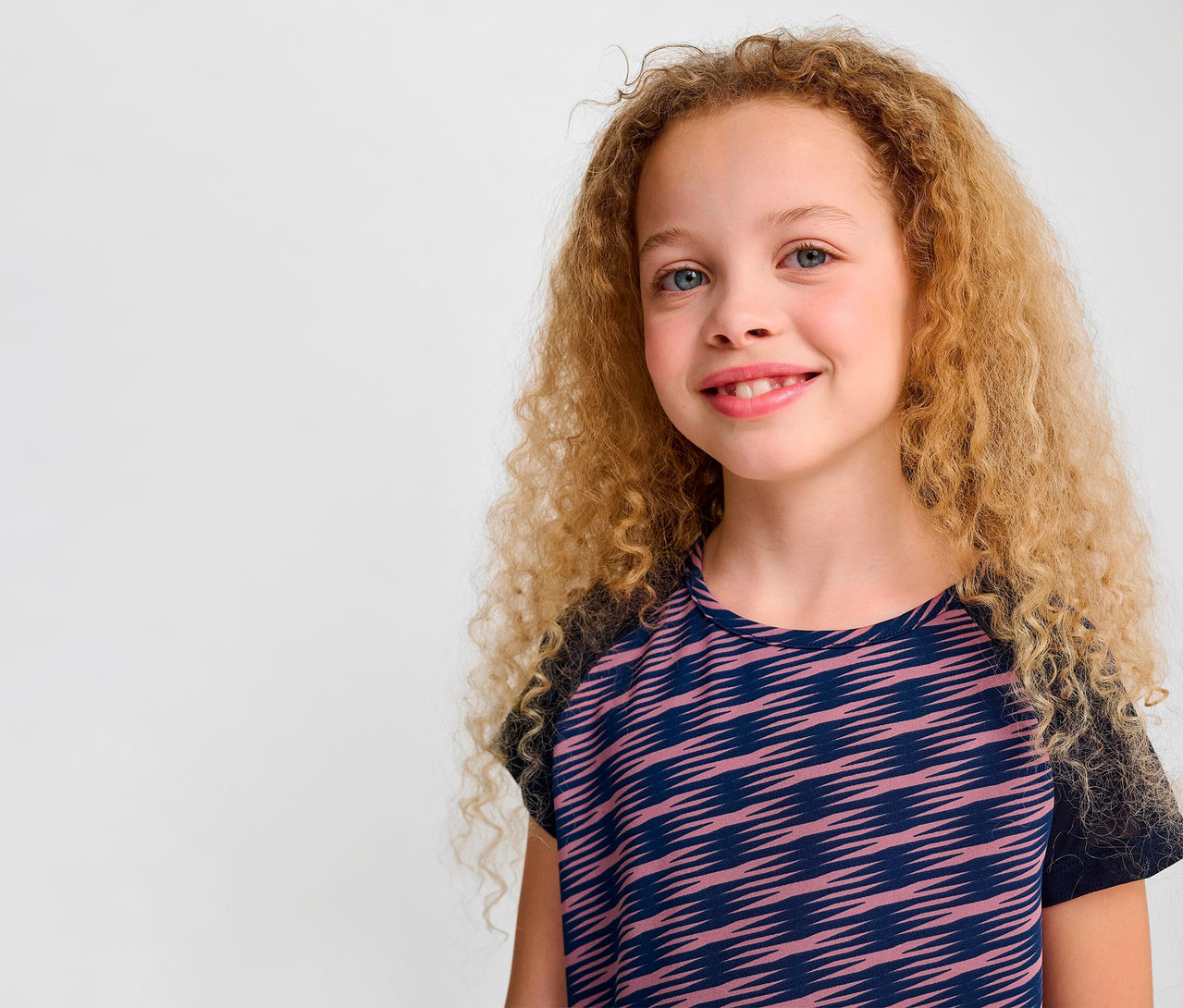 Portrait d'une fillette souriante aux cheveux blonds bouclés portant un t-shirt S/S HUMMEL HMLJR Mix.