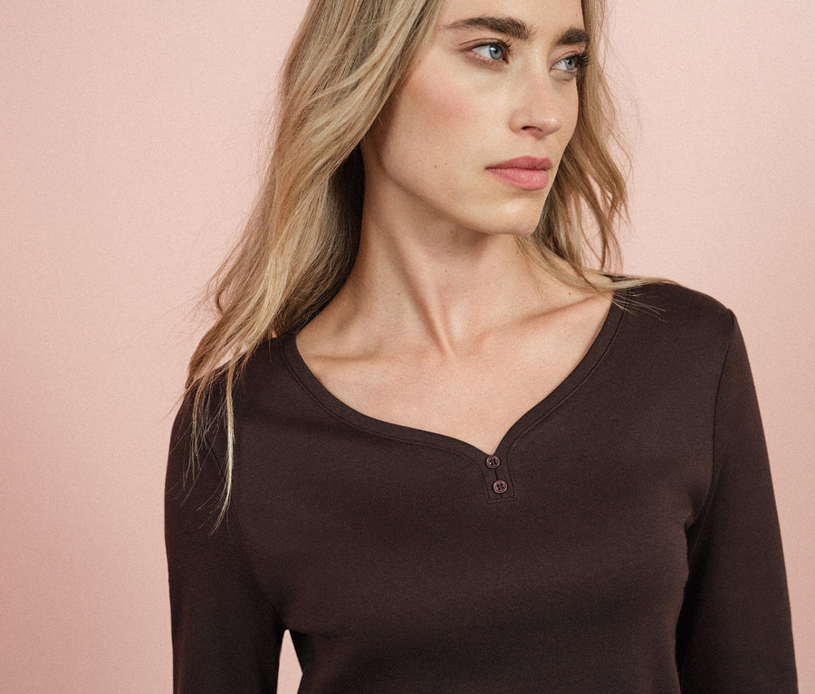 Une femme blonde aux yeux bleus porte un t-shirt marron à manches longues.